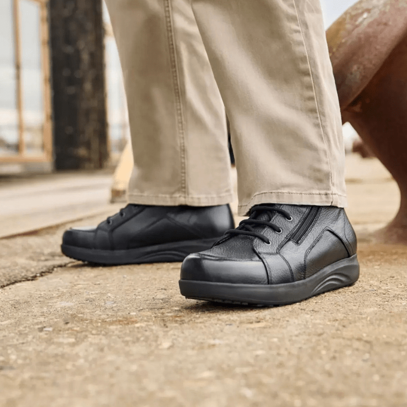 Um homem com calças bege e botas New Feet pretas de homem em pé numa superfície cinzenta