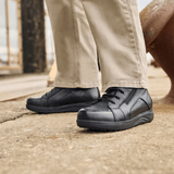 Um homem com calças bege e botas New Feet pretas de homem em pé numa superfície cinzenta